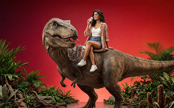 Cinematic scene — woman on a T-Rex with tropical foliage and bold red backdrop
